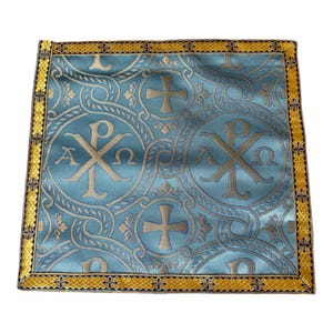 Könnte beinhalten: Ein quadratisches hellblaues Stofftuch mit einem sich wiederholenden goldfarbenen Muster aus Chi-Rho-Symbolen, Alpha- und Omega-Buchstaben sowie Kreuzen. Die Kanten sind mit einer breiten, dekorativen gold- und dunkelblauen Webkante versehen.