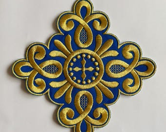 May include: A blue and gold embroidered applique with a symmetrical design. The applique features a central cross and a repeating pattern of swirls and curves. The gold embroidery is detailed and stands out against the blue background.