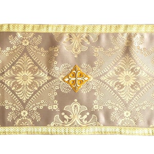 以下が含まれることがあります： 金と茶色のテーブルランナーで、金色の刺繍模様とフリンジが付いています。ランナーの中央には、十字架のあるダイヤモンド型のモチーフがあります。