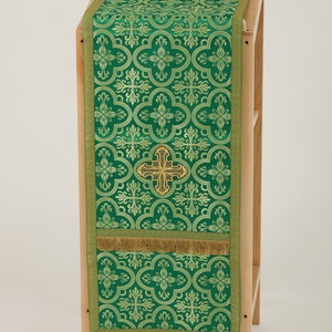 May include: Green altar runner with a gold cross and a repeating pattern of gold quatrefoils. The runner is on a wooden stand.