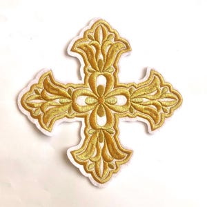 May include: Gold embroidered cross patch with intricate floral patterns and a white border. The cross has a symmetrical design with a central flower-like motif and stylized flourishes. Suitable for adding a decorative touch to clothing or accessories.