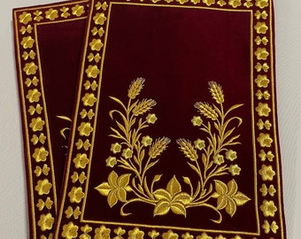 Puede incluir: Dos manteles individuales de terciopelo burdeos con diseños florales bordados en oro y un borde dorado.