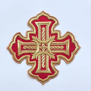 May include: Embroidered red cross with gold trim and detailing. The cross features a decorative design with intricate patterns and a rope-like texture in the center. The cross is centered on a white background.