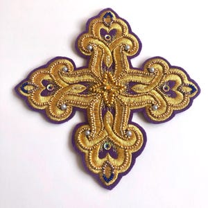 May include: A cross-shaped patch featuring intricate gold metallic thread embroidery on a purple felt base. The design is embellished with small silver-toned beads, iridescent sequins, and tiny blue seed beads arranged in a symmetrical pattern.