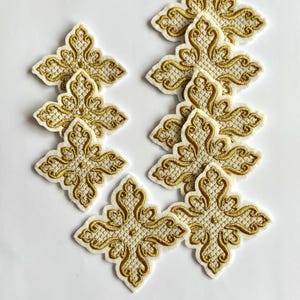 May include: A collection of embroidered appliques in a cross-like shape. The appliques feature a cream-colored base with intricate gold-colored embroidery. The design is symmetrical and ornate, suitable for embellishing garments or crafts.