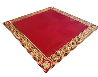 Puede incluir: Un textil rojo cuadrado con un borde dorado adornado. El borde dorado presenta un diseño intrincado y repetitivo con elementos florales y geométricos. Las esquinas tienen emblemas dorados decorativos. El textil probablemente se utiliza con fines decorativos.