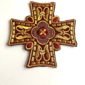 Op de afbeelding: Een rood geborduurd kruis met gouden draaddetails. Het kruis heeft een centraal bloemmotief met een gouden X in het midden.