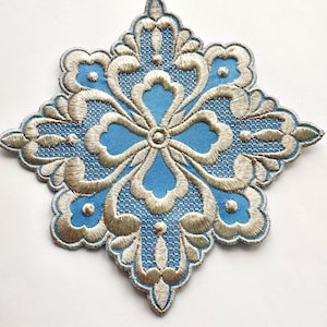 May include: A detailed, embroidered patch with a symmetrical floral design. The patch features a light blue center with gold-colored embroidery outlining the floral pattern. The design is a four-pointed star shape.