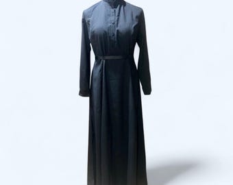 May include: A long, black dress with long sleeves and a high collar. The dress features a button-down front and a thin belt at the waist. The dress is displayed on a mannequin against a white background. The dress is a solid color and appears to be made of a soft fabric.