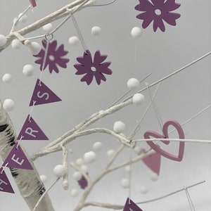 Op de afbeelding: Een witte berkentakboom versierd met paarse en roze viltbloemen, harten en driehoekige vlaggetjes met het woord "HERS" erop. De boom is ook versierd met witte pompoenen.