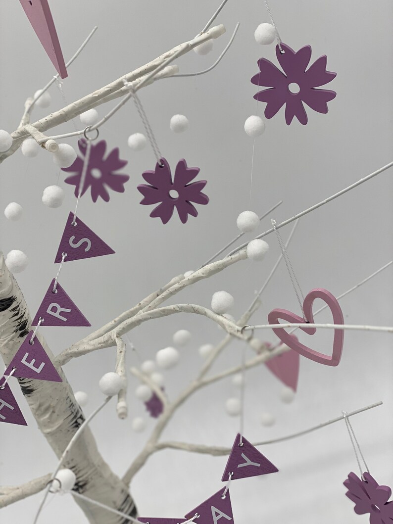 Op de afbeelding: Een witte berkentakboom versierd met paarse en roze viltbloemen, harten en driehoekige vlaggetjes met het woord "HERS" erop. De boom is ook versierd met witte pompoenen.