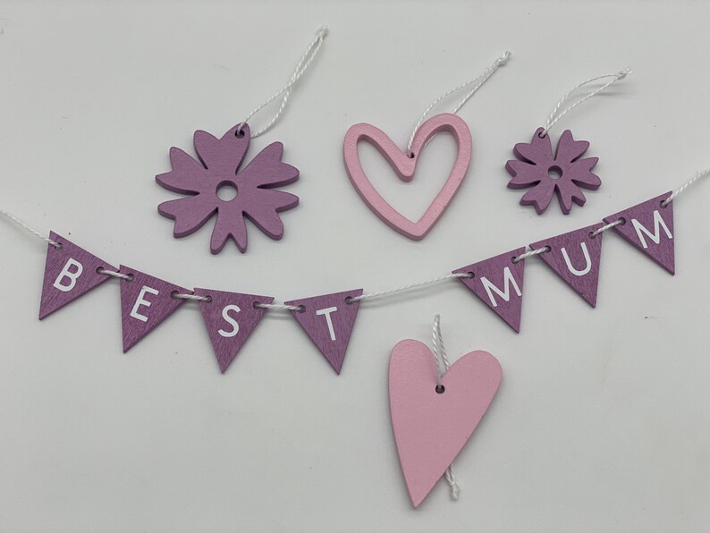 Op de afbeelding: Een paarse en roze houten banner met de woorden "BEST MUM" geregen aan een wit touwtje. De banner is versierd met twee paarse bloemen en twee roze harten.