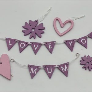 Op de afbeelding: Een set van paarse en roze houten decoraties met een reeks vlaggen waarop "LOVE YOU MUM" staat. De vlaggen zijn aan elkaar geregen met wit touw en hebben roze harten en bloemen.