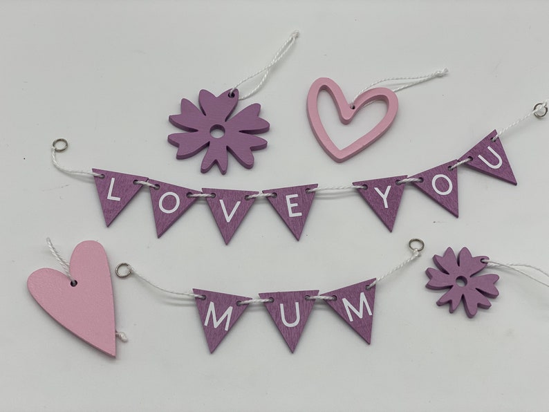 Op de afbeelding: Een set van paarse en roze houten decoraties met een reeks vlaggen waarop "LOVE YOU MUM" staat. De vlaggen zijn aan elkaar geregen met wit touw en hebben roze harten en bloemen.