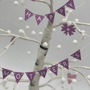 Op de afbeelding: Een witte berkenboom versierd met paarse en witte vlaggetjes waarop "Happy Mothers" staat. De vlaggetjes zijn versierd met witte pompoentjes. Er zijn ook twee roze harten en een paarse bloem op de boom.