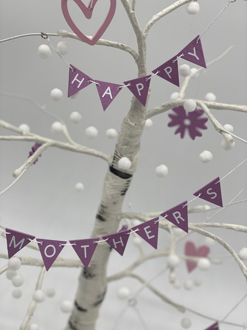 Op de afbeelding: Een witte berkenboom versierd met paarse en witte vlaggetjes waarop "Happy Mothers" staat. De vlaggetjes zijn versierd met witte pompoentjes. Er zijn ook twee roze harten en een paarse bloem op de boom.