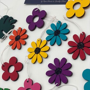 May include: A set of colourful wooden flower decorations with glitter centres. The decorations are strung together with white twine and are ideal for adding a touch of whimsy to any space.