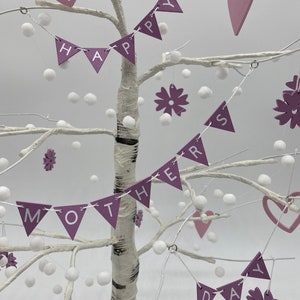 Op de afbeelding: Een witte berkenboom versierd met paarse en witte decoraties. De boom is versierd met een banner met de tekst "Happy Mother's Day" en met harten, bloemen en pompoenen.