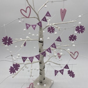Op de afbeelding: Een witte berkenboom versierd met roze en witte ornamenten. De ornamenten spellen "Happy Mothers Day" met harten en bloemen.
