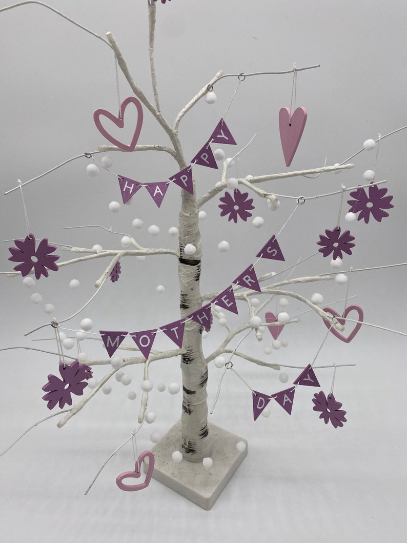 Op de afbeelding: Een witte berkenboom versierd met roze en witte ornamenten. De ornamenten spellen "Happy Mothers Day" met harten en bloemen.