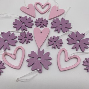 Op de afbeelding: Een set van 14 houten ornamenten in roze en paarse tinten. De ornamenten zijn in de vorm van harten en bloemen. Ze hebben een rustieke, handgemaakte uitstraling en zijn perfect om elke ruimte een vleugje whimsy te geven.