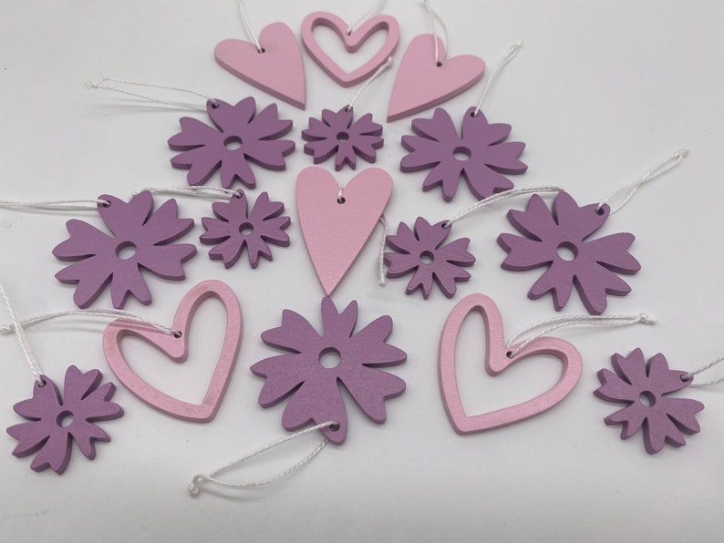 Op de afbeelding: Een set van 14 houten ornamenten in roze en paarse tinten. De ornamenten zijn in de vorm van harten en bloemen. Ze hebben een rustieke, handgemaakte uitstraling en zijn perfect om elke ruimte een vleugje whimsy te geven.