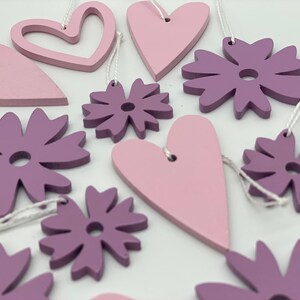 Op de afbeelding: Een set van 10 houten ornamenten in roze en paarse tinten. De ornamenten zijn in de vorm van harten en bloemen. Ze zijn aan een wit touwtje geregen en zouden perfect zijn voor een kinderkamer of een kinderkamer.