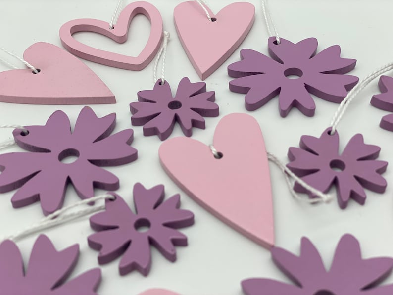 Op de afbeelding: Een set van 10 houten ornamenten in roze en paarse tinten. De ornamenten zijn in de vorm van harten en bloemen. Ze zijn aan een wit touwtje geregen en zouden perfect zijn voor een kinderkamer of een kinderkamer.