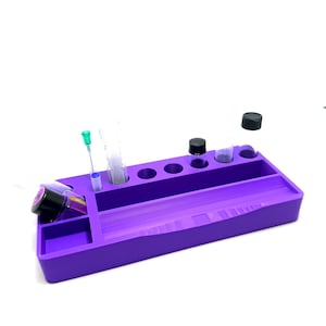 Puede incluir: Una bandeja organizadora morada con varios compartimentos y agujeros. Contiene viales, jeringas y botellas con tapas negras. La bandeja est&aacute; dise&ntilde;ada para mantener los objetos peque&ntilde;os organizados y accesibles. La bandeja es de material s&oacute;lido.