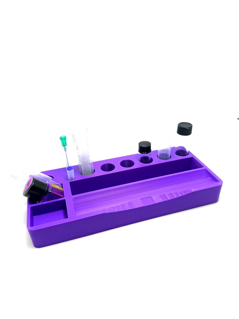 Puede incluir: Una bandeja organizadora morada con varios compartimentos y agujeros. Contiene viales, jeringas y botellas con tapas negras. La bandeja est&aacute; dise&ntilde;ada para mantener los objetos peque&ntilde;os organizados y accesibles. La bandeja es de material s&oacute;lido.