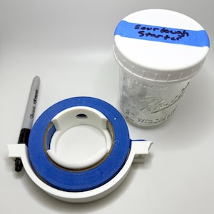 May include: A clear glass jar with a white lid, labeled "Sourdough Starter" with blue tape. A roll of blue painter's tape sits in a white dispenser, and a black Sharpie marker is nearby. The items are on a white surface.