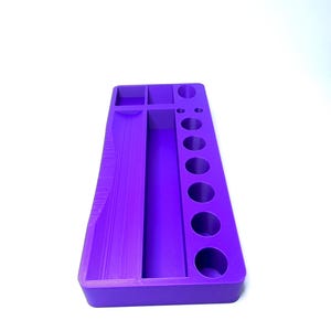 Puede incluir: Un organizador morado con varios compartimentos y agujeros circulares. La bandeja rectangular tiene una secci&oacute;n larga y estrecha, varios compartimentos cuadrados y rectangulares peque&ntilde;os y una serie de siete agujeros circulares de varios tama&ntilde;os. El art&iacute;culo est&aacute; hecho de un material s&oacute;lido y liso.