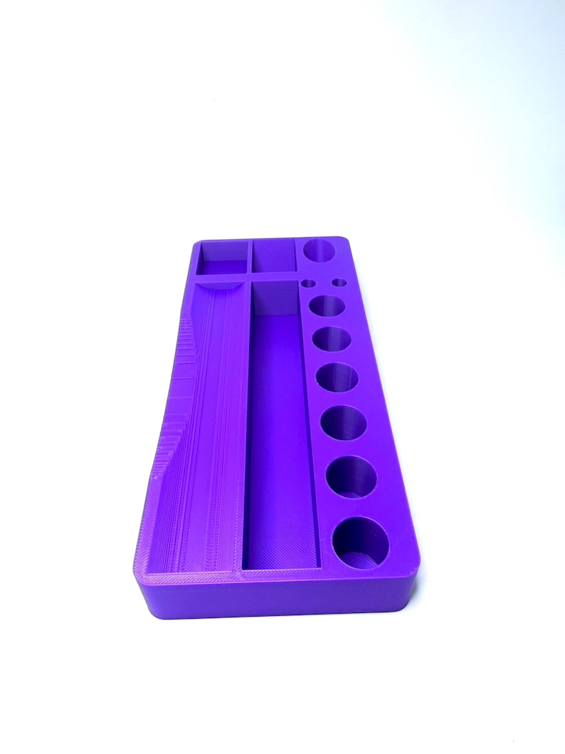 Puede incluir: Un organizador morado con varios compartimentos y agujeros circulares. La bandeja rectangular tiene una secci&oacute;n larga y estrecha, varios compartimentos cuadrados y rectangulares peque&ntilde;os y una serie de siete agujeros circulares de varios tama&ntilde;os. El art&iacute;culo est&aacute; hecho de un material s&oacute;lido y liso.