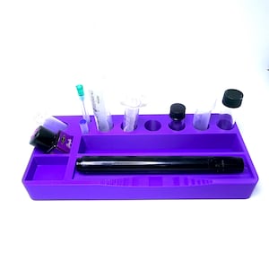 Puede incluir: Una bandeja organizadora morada con varios compartimentos y soportes. Contiene un bol&iacute;grafo negro, peque&ntilde;os viales, jeringas y frascos de tinta. La bandeja est&aacute; dise&ntilde;ada para guardar objetos peque&ntilde;os y herramientas.