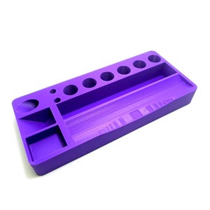 Puede incluir: Un organizador de escritorio rectangular morado con varios compartimentos y agujeros circulares. El organizador tiene un largo canal central, varios agujeros circulares peque&ntilde;os y compartimentos rectangulares y circulares m&aacute;s peque&ntilde;os.