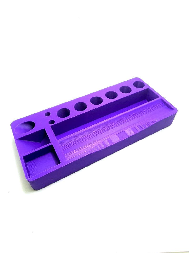 Puede incluir: Un organizador de escritorio rectangular morado con varios compartimentos y agujeros circulares. El organizador tiene un largo canal central, varios agujeros circulares peque&ntilde;os y compartimentos rectangulares y circulares m&aacute;s peque&ntilde;os.