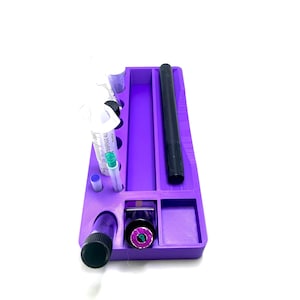Puede incluir: Una bandeja organizadora morada con varios compartimentos y art&iacute;culos. Incluye jeringas, un bol&iacute;grafo negro y peque&ntilde;os recipientes. La bandeja es rectangular y est&aacute; sobre una superficie blanca.