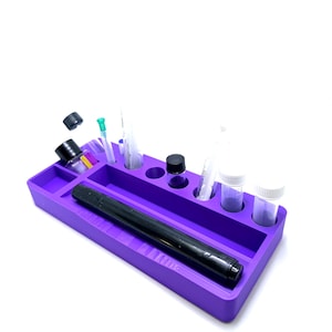Puede incluir: Una bandeja de silicona morada con varios compartimentos y un marcador negro. La bandeja contiene peque&ntilde;os viales, jeringas y botellas. La bandeja est&aacute; dise&ntilde;ada para organizar objetos peque&ntilde;os. La bandeja est&aacute; sobre un fondo blanco.
