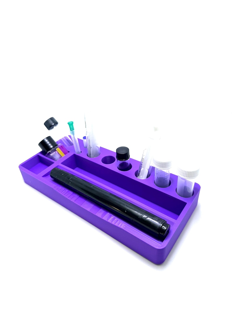 Puede incluir: Una bandeja de silicona morada con varios compartimentos y un marcador negro. La bandeja contiene peque&ntilde;os viales, jeringas y botellas. La bandeja est&aacute; dise&ntilde;ada para organizar objetos peque&ntilde;os. La bandeja est&aacute; sobre un fondo blanco.