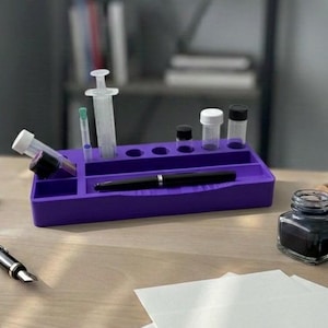 Puede incluir: Un organizador de escritorio morado contiene una jeringa, viales y un bol&iacute;grafo negro. Un peque&ntilde;o tintero y una pila de papel blanco est&aacute;n sobre el escritorio de madera. El fondo incluye una estanter&iacute;a.
