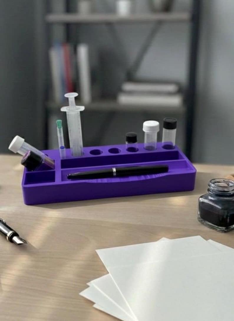 Puede incluir: Un organizador de escritorio morado contiene una jeringa, viales y un bol&iacute;grafo negro. Un peque&ntilde;o tintero y una pila de papel blanco est&aacute;n sobre el escritorio de madera. El fondo incluye una estanter&iacute;a.