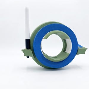 May include: A sage green plastic tape dispenser holding a roll of bright blue painter's tape. The dispenser features a vertical slot on the left side containing a black and white marker. The design is circular with a flat base for stability.
