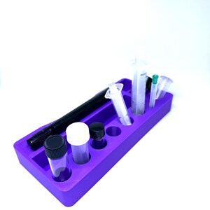 Puede incluir: Una bandeja organizadora morada que contiene varios suministros de laboratorio. La bandeja contiene viales transparentes con tapas negras y blancas, jeringas, un marcador negro y otros objetos peque&ntilde;os. Los art&iacute;culos est&aacute;n ordenados.