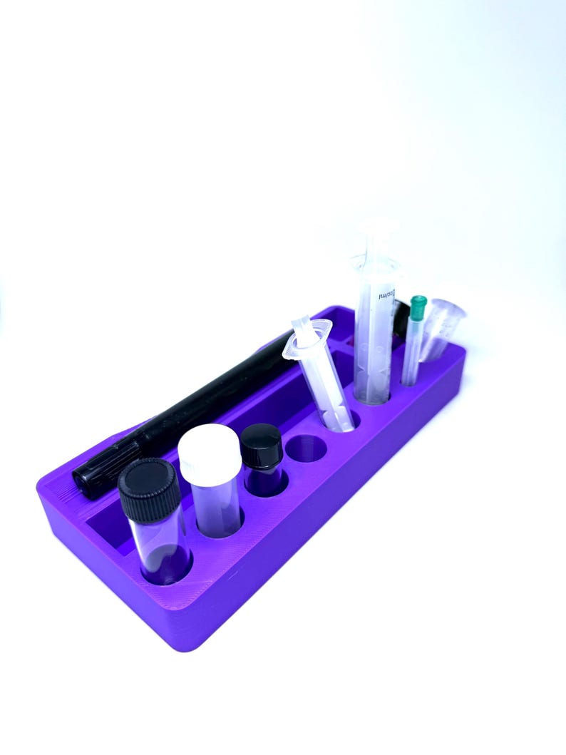 Puede incluir: Una bandeja organizadora morada que contiene varios suministros de laboratorio. La bandeja contiene viales transparentes con tapas negras y blancas, jeringas, un marcador negro y otros objetos peque&ntilde;os. Los art&iacute;culos est&aacute;n ordenados.