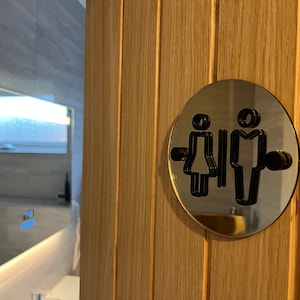 May include: A round, mirrored sign with black stick figure icons for a restroom. The sign is mounted on a wooden door.