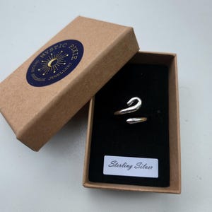 Könnte beinhalten: Ein silberfarbener Ring mit geschwungenem Design in einer offenen braunen Schachtel mit schwarzem Innenraum. Die Schachtel hat einen blauen Aufkleber mit dem Text "MYSTIC PIXIE HANDMADE JEWELLERY". Ein weißes Etikett in der Schachtel lautet "Sterling Silver".