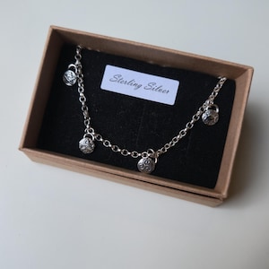 May include: A sterling silver necklace with multiple decorative charms, presented in a small brown box with a black interior. The charms have a detailed, textured design. The box contains a label that reads "Sterling Silver".