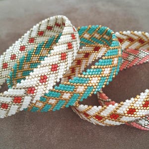 Peut inclure: Trois bracelets en perles avec des motifs géométriques en blanc, turquoise et rouge corail. Les bracelets sont tissés avec de petites perles et ont un fil doré qui les traverse.