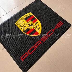 Puede incluir: Felpudo negro con un logotipo de Porsche amarillo y rojo y la palabra "PORSCHE" en letras rojas.