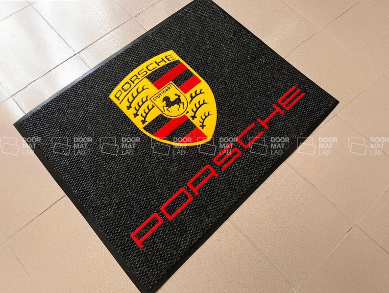 Puede incluir: Felpudo negro con un logotipo de Porsche amarillo y rojo y la palabra "PORSCHE" en letras rojas.